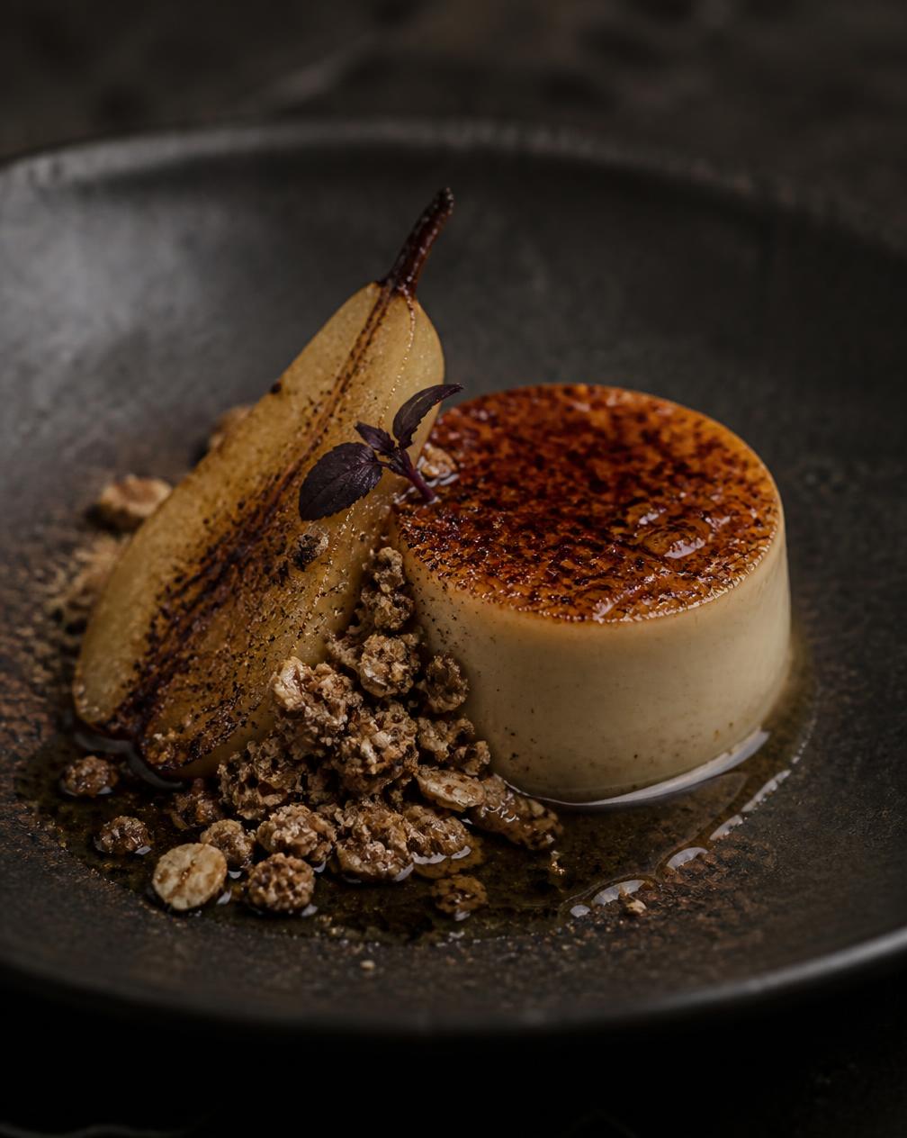 Burnt honey custard with charred pear and oat crumble in a dark ceramic bowl